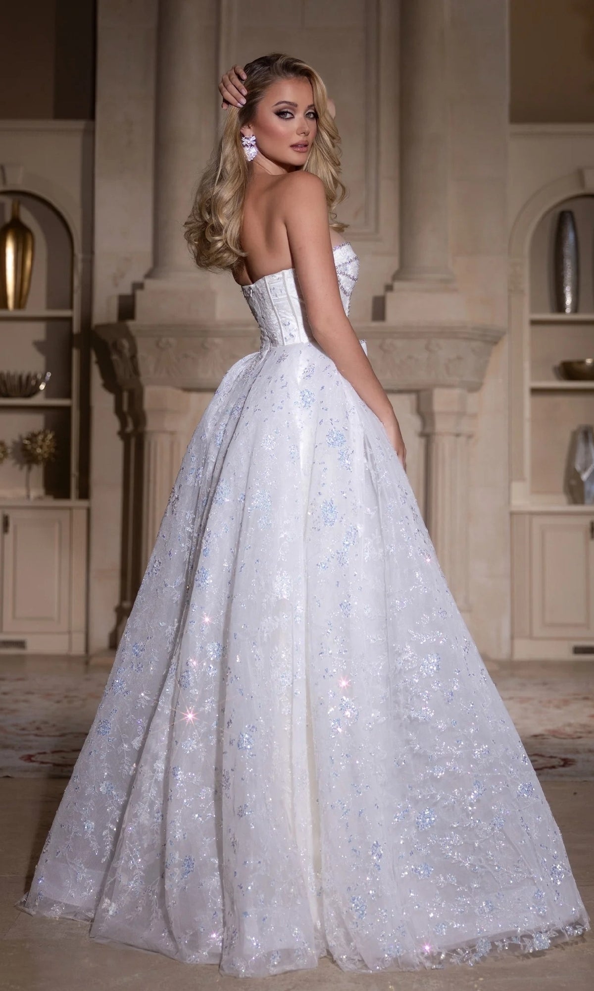 A woman in the Portia and Scarlett PS25315 long prom dress, featuring a strapless corset bodice with glittery details, stands indoors and looks over her shoulder.